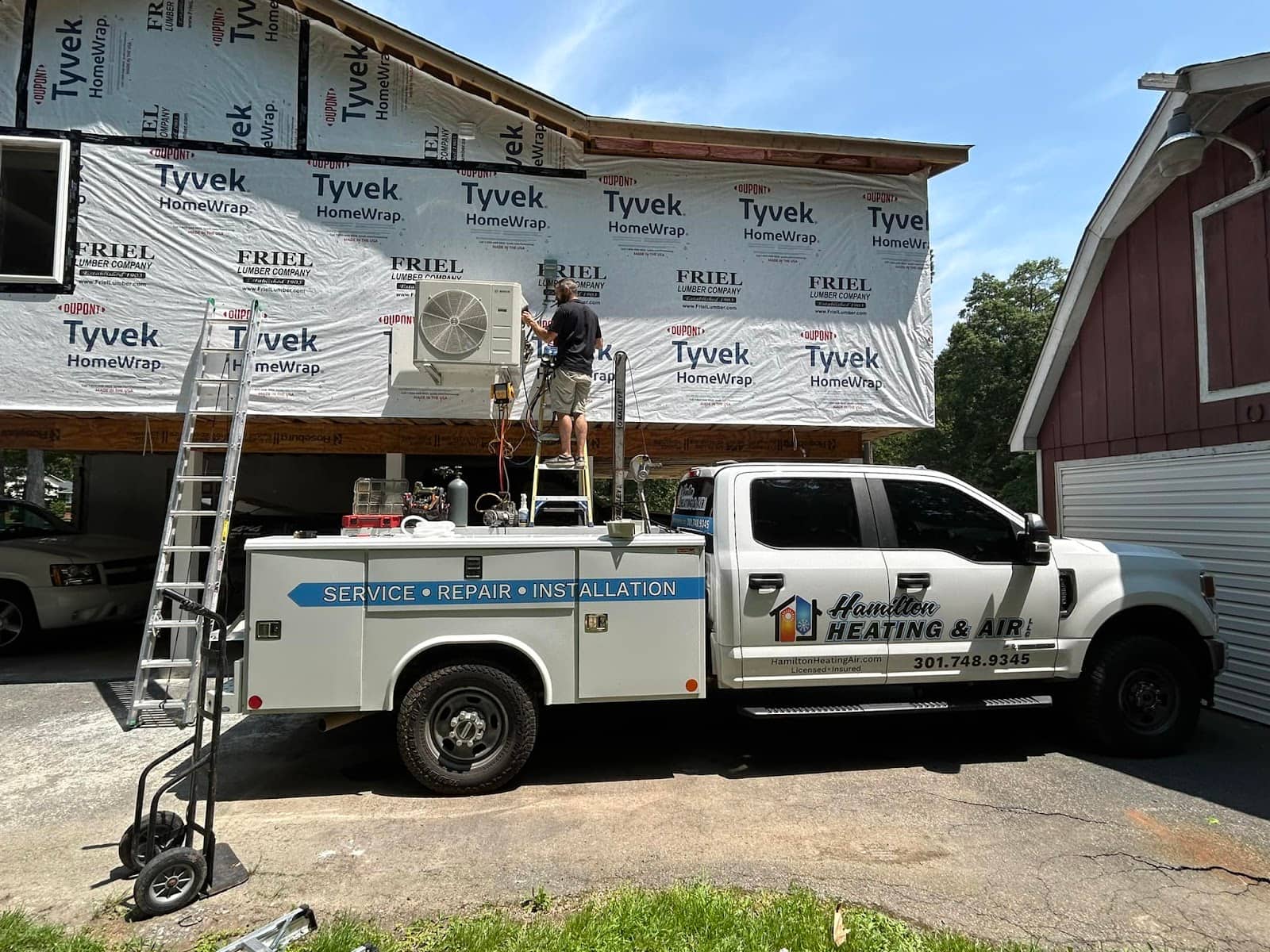Hamilton technician on a ladder mounting a ductless unit, with the lettered service truck in the drive