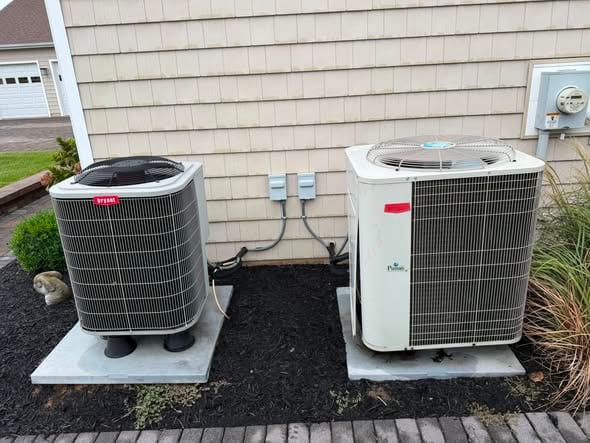 Two outdoor units set on pads beside a tan sided home with black mulch