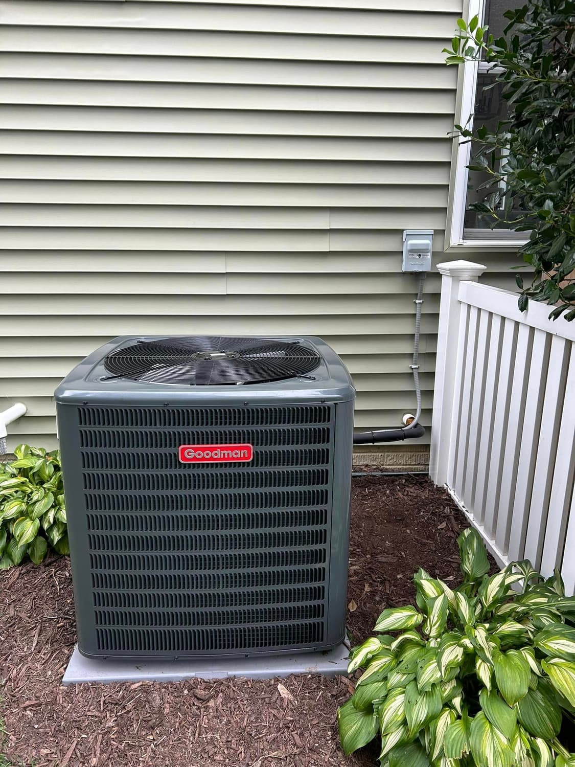 New outdoor condenser on a fresh pad beside a white vinyl fence and hostas